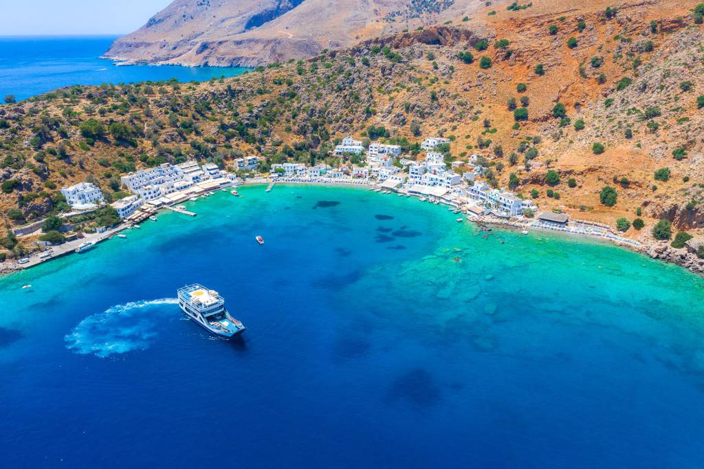 Aerial view of Loutro Seaside Village with its whitewashed little houses, surrounded by mountains and a ship leaving the village.