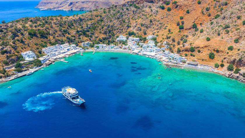 Aerial view of Loutro Seaside Village with its whitewashed little houses, surrounded by mountains and a ship leaving the village.