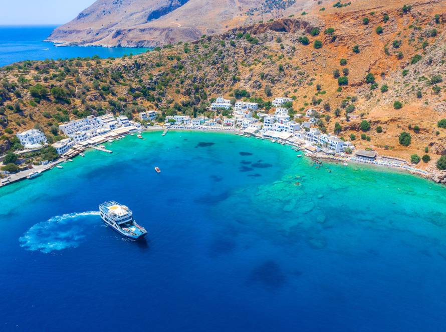 Aerial view of Loutro Seaside Village with its whitewashed little houses, surrounded by mountains and a ship leaving the village.