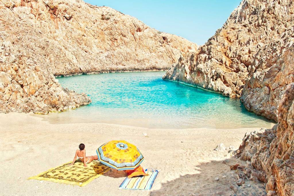 A woman sits on a towel below her umbrella in Seitan Limania Beach.