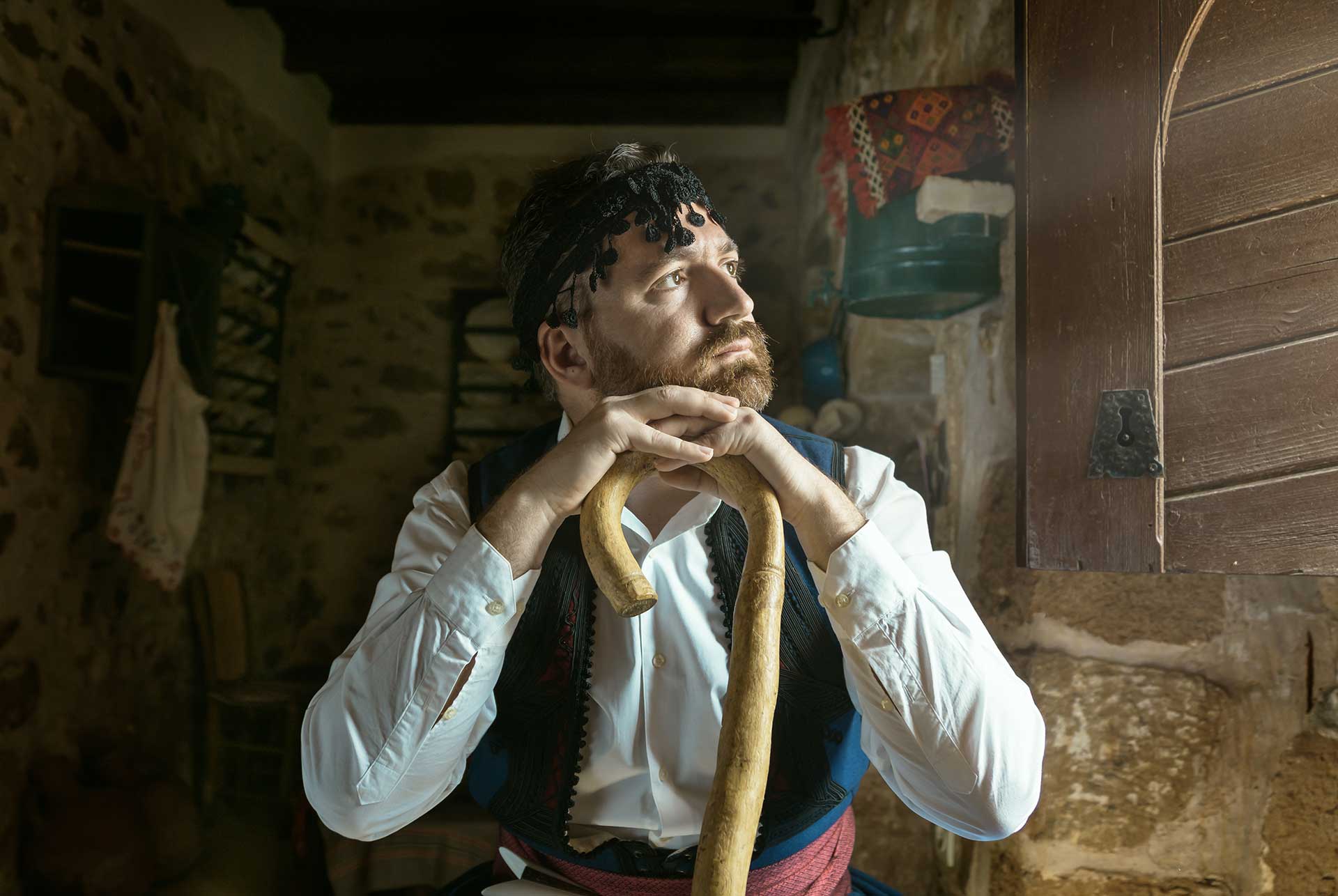 A young man dressed in Cretan traditional clothing, wearing a sariki on his head and holding a katsouna walking stick.