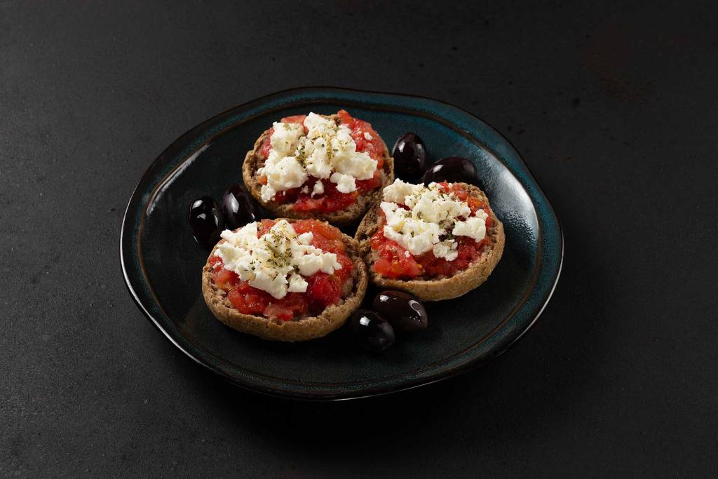 Cretan Dakos with rusk, tomato, mizithra cheese, olives, and oregano served on a black plate.