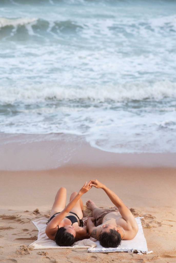 A young couple is lying on the beach, holding hands.