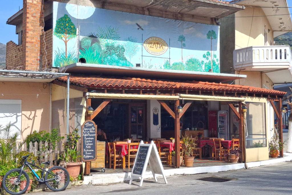 The entrance and blue sign of Platia Cafe Tavern in Kandanos Village (South Chania) with its wooden tables and chairs.