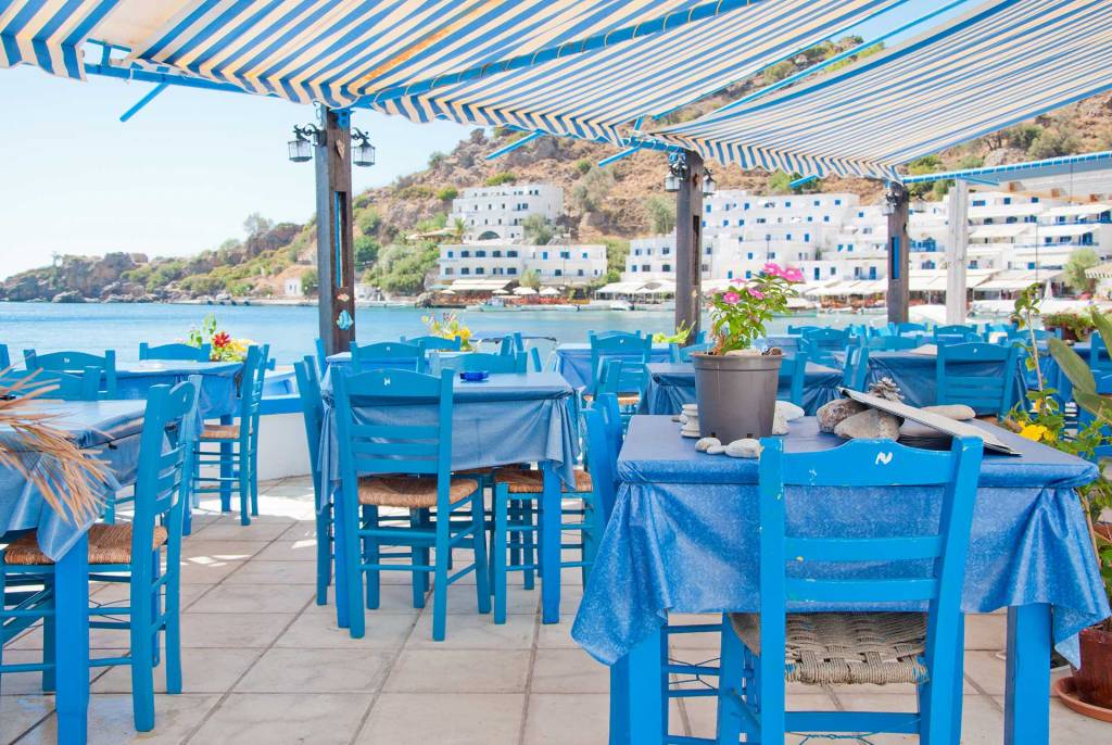 A picturesque little tavern with blue chairs and tables in Loutro Village offering beautiful sea and village views.