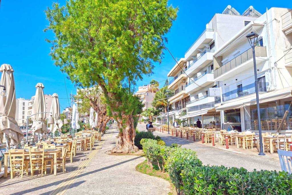 The tables, chairs, and umbrellas of picturesque seaside restaurants in Nea Chora (Chania), surrounded by trees, and the local houses.