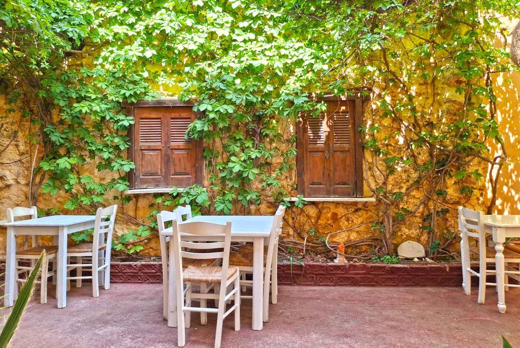 The white tables and chairs of a picturesque restaurant in Chania Town, surrounded by vegetation and yellow walls.
