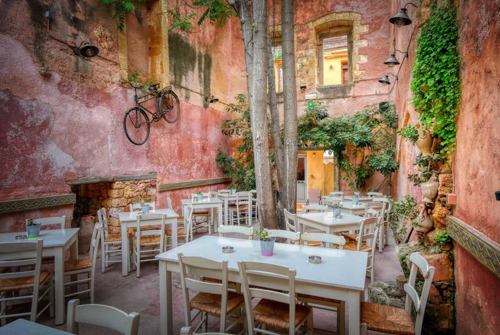 The white tables and chairs of the picturesque Mesostrato Restaurant in Chania Old Town surrounded by beautiful vegetation and a hanging bike as part of its decoration.