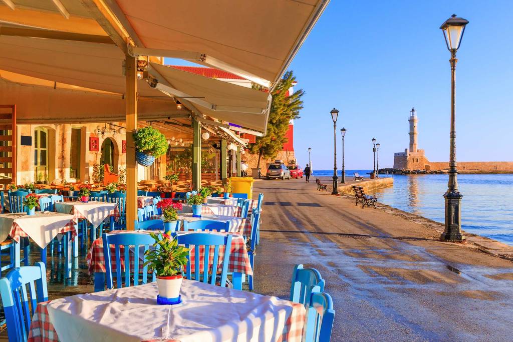 The tables and chairs of a restaurant in Chania's Venetian Old Harbor offering views of the Egyptian Lighthouse and the sea.