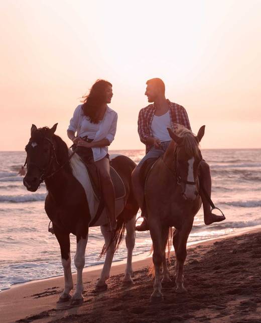 A couple doing horseback riding on a sandy beach during sunset.
