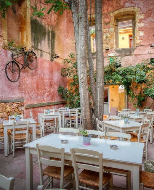 The white tables and chairs of the picturesque Mesostrato Restaurant in Chania Old Town surrounded by beautiful vegetation and a hanging bike as part of its decoration.