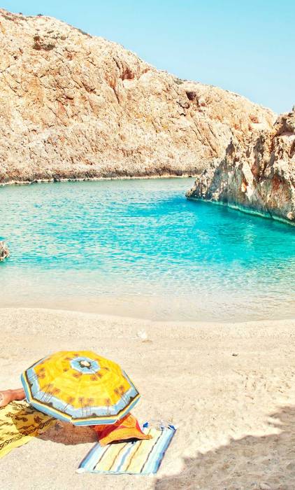 A woman sits on a towel below her umbrella in Seitan Limania Beach.